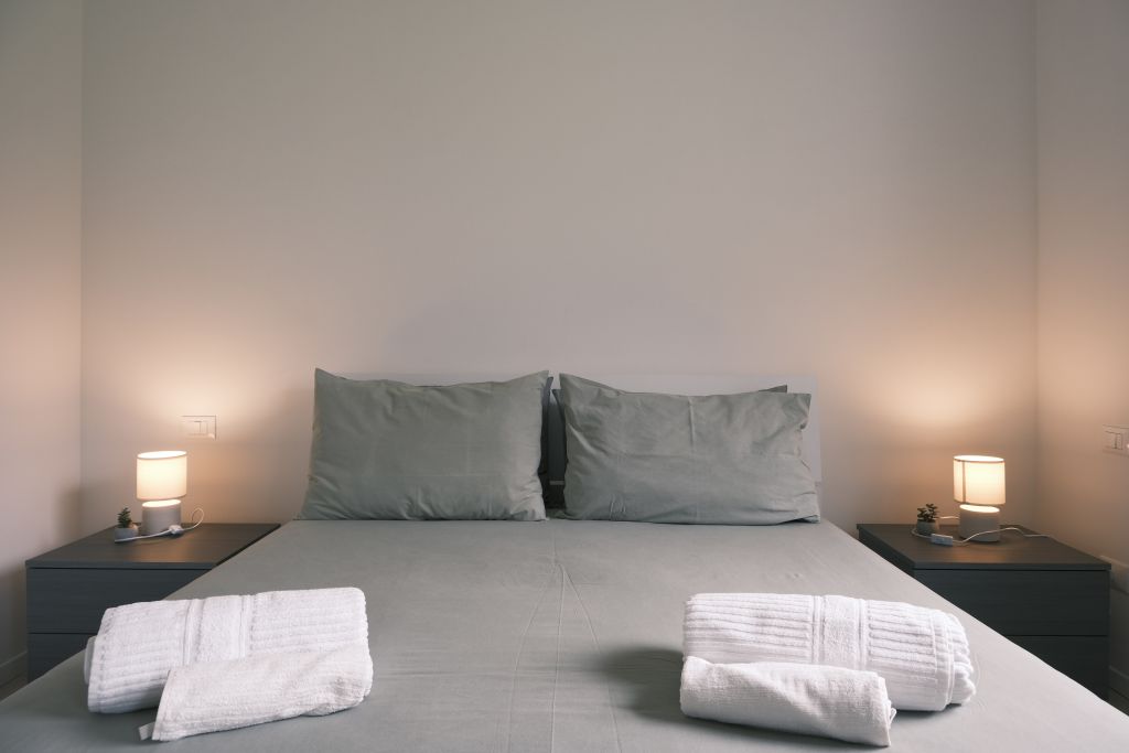 Letto matrimoniale con cuscini grigi, due lampade da comodino accese e asciugamani bianchi piegati