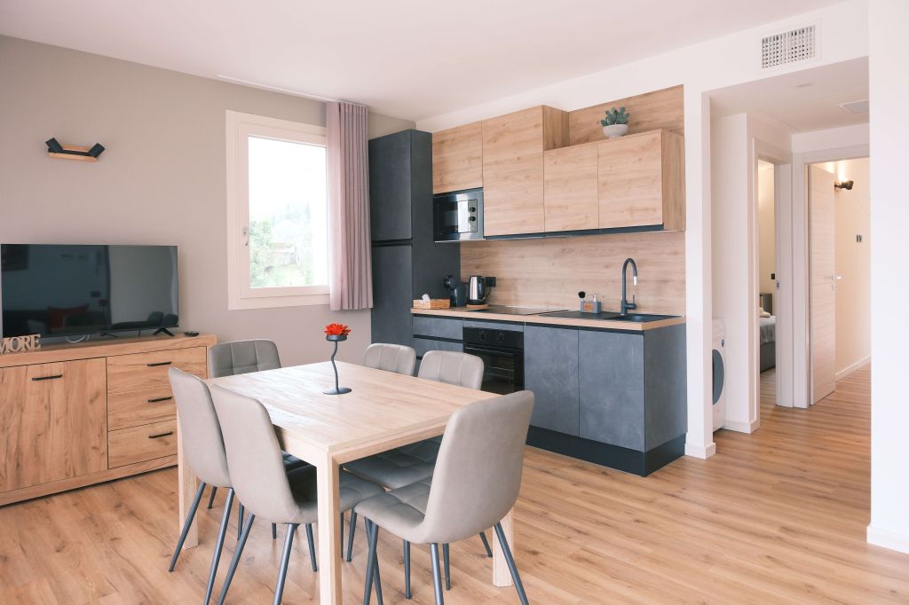 Cucina moderna con mobili in legno chiaro, tavolo con sedie grigie e finestra luminosa.