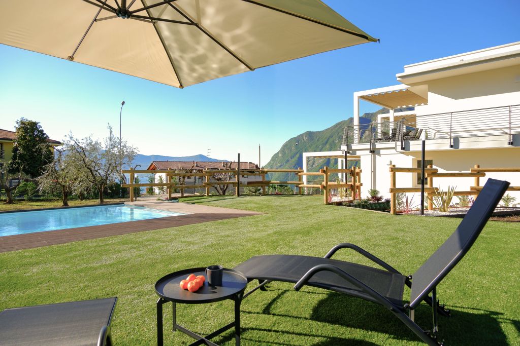 Terrazza con piscina, ombrellone, sdraio e tavolino con frutta e tazza, vista montagne e cielo sereno