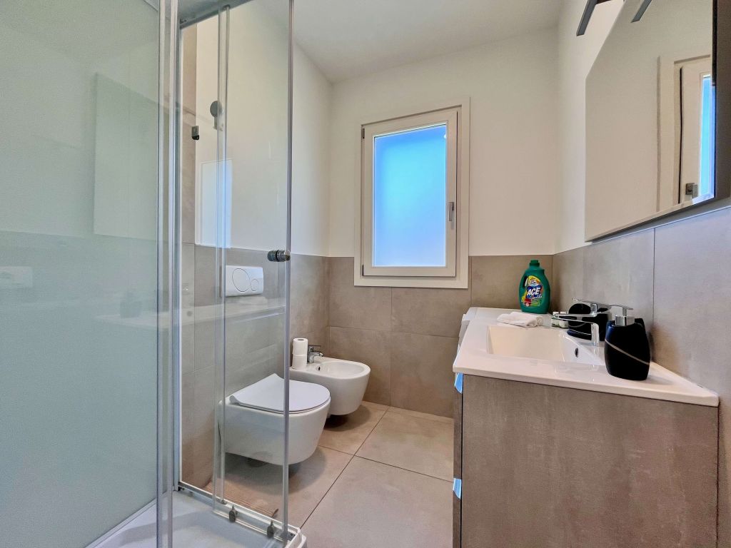 Bagno moderno con doccia in vetro, wc, bidet e lavabo con specchio e finestra.