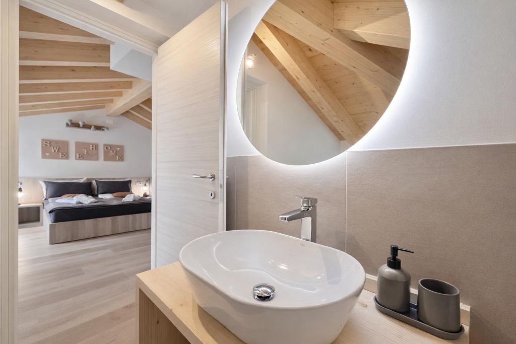 Lavabo moderno con specchio rotondo illuminato e vista parziale di camera da letto con travi in legno