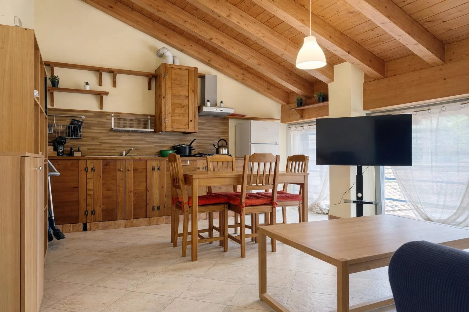 Cucina in legno con travi a vista, tavolo con sedie e TV vicino a grandi finestre con tende bianche.
