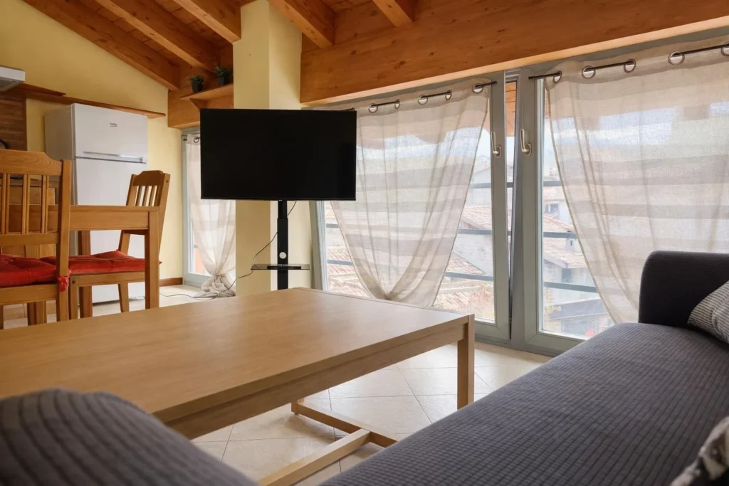 Soggiorno con divano grigio, tavolino in legno, TV e finestre con tende leggere.
