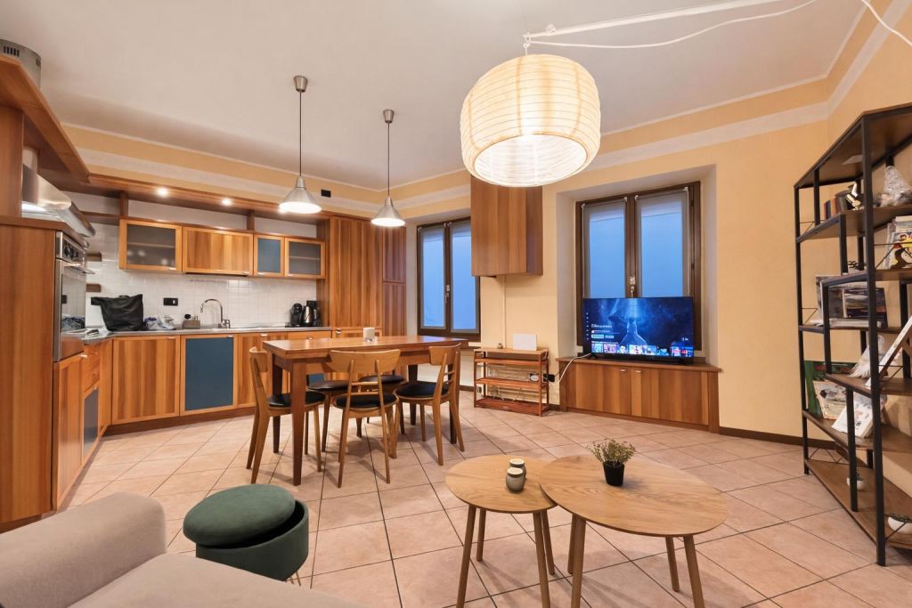 Soggiorno con cucina a vista, tavolo in legno, TV e libreria metallica