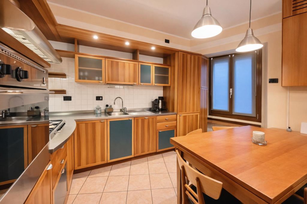 Cucina con mobili in legno e acciaio, tavolo con sedie e finestra con tende.