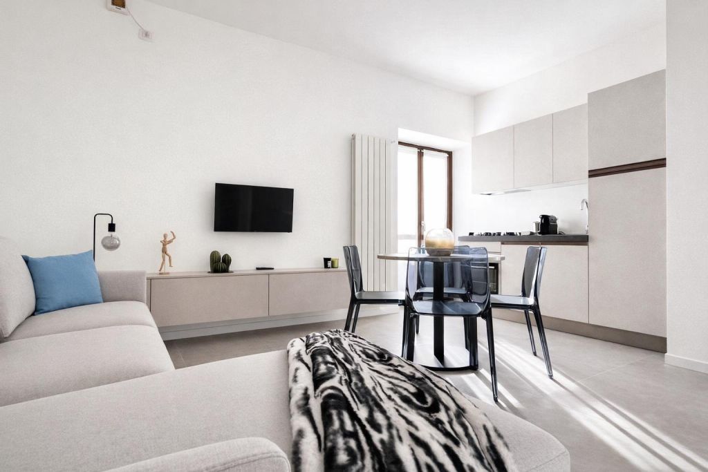 Soggiorno luminoso con divano beige, tavolo con sedie trasparenti e cucina moderna.