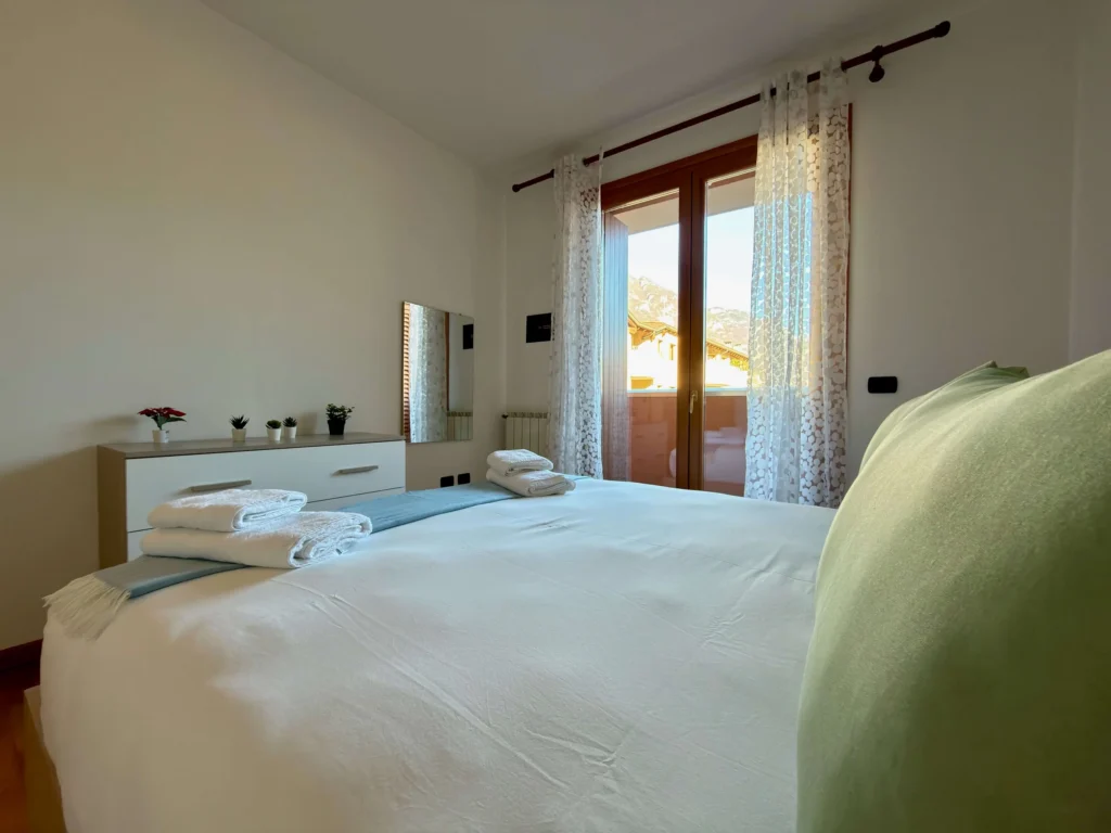 Camera con letto matrimoniale, cassettiere, specchio e porta finestra con tende bianche