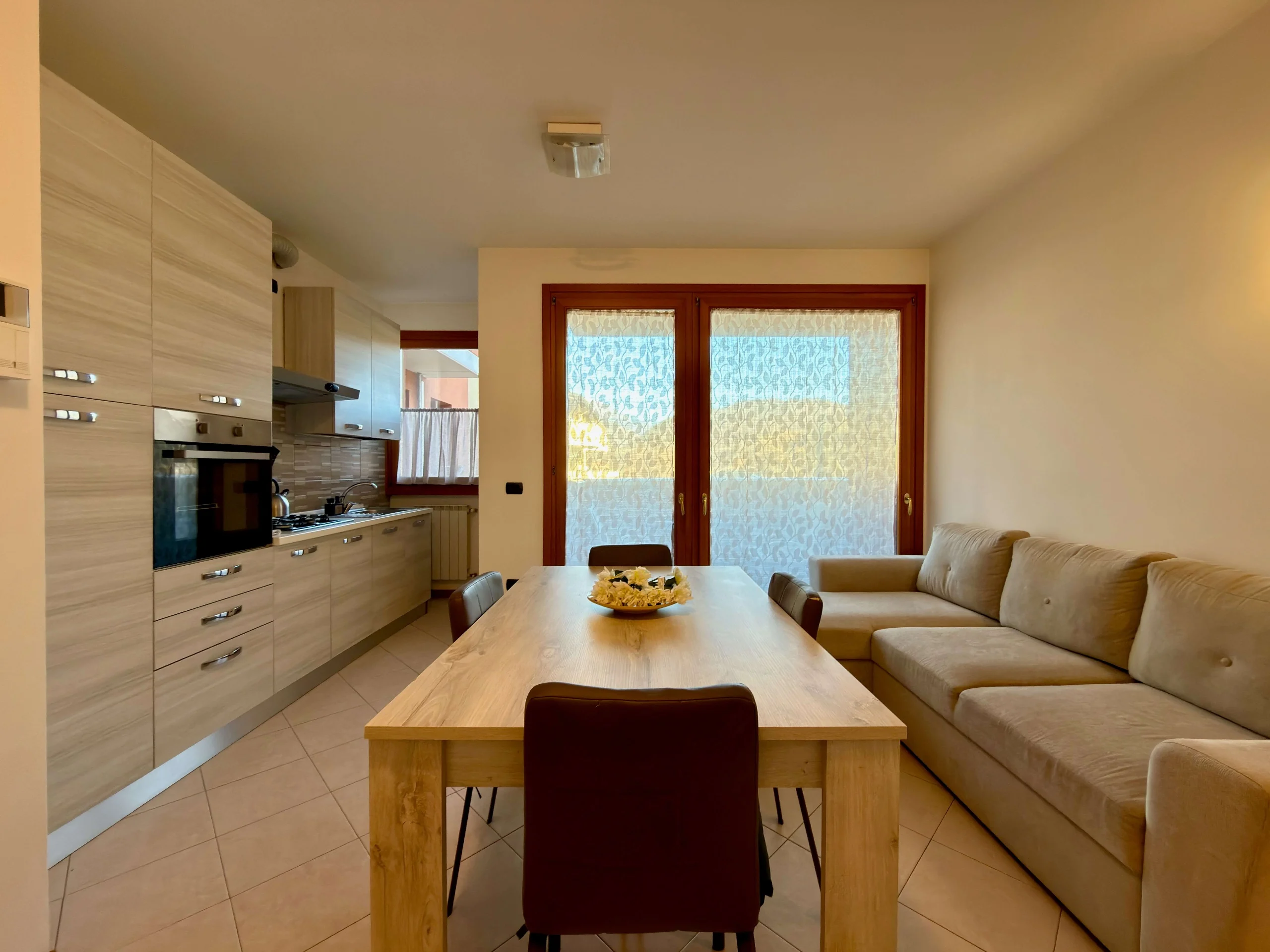 Zona giorno con cucina moderna, tavolo in legno e divano beige vicino a finestra con tende
